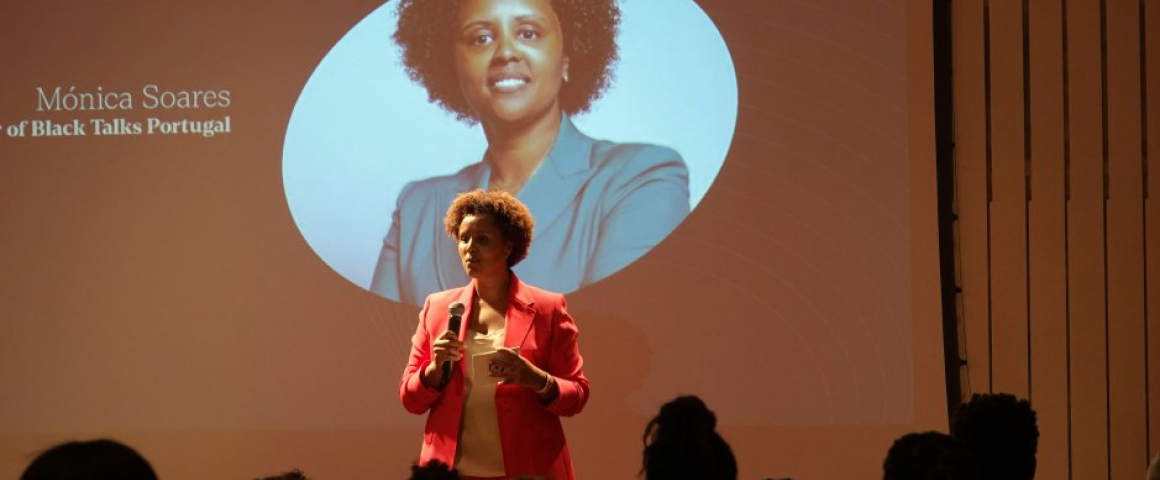A imagem mostra uma mulher a segurar um microfone. Ela veste um blazer rosa e está em frente a um ecrã de projeção. No ecrã, aparece uma fotografia dela em plano médio, com o nome "Mónica Soares" e a descrição abaixo: "Founder of Black Talks Portugal". O público está parcialmente visível na parte inferior da imagem, destacando-se como silhuetas contra a luz projetada. O cenário é de uma apresentação ou palestra.